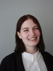 Profilbild von Matilda Bieder. Sie hat dunkle schulterlange Haare, steht vor einer grauen Wand und lächelt freundlich.