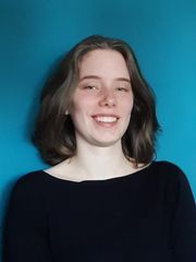 Profil Bild von Stine Behrmann. Sie hat braune schulterlange Haare, steht vor einem Blauen Hintergrund und hat ein breites, freundliches Lächeln