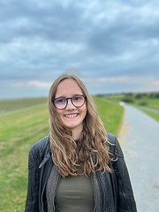 Emily hat lange blonde Haare und trägt eine Brille. Sie steht lächelnd auf einem Deich.